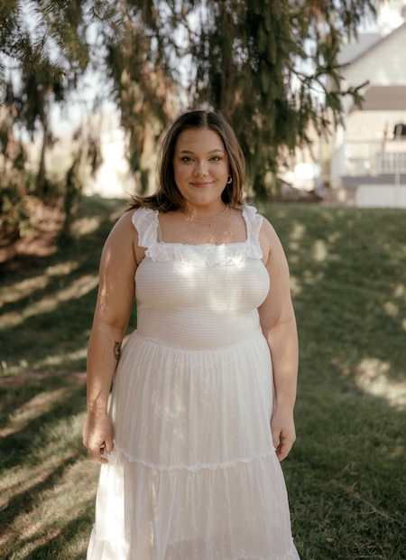 Leslie Harker wearing white dress standing under evergreen tree